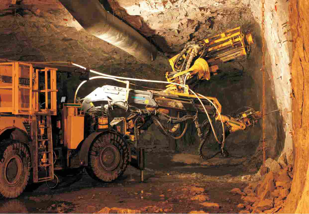 Photo of a tractor-like vehicle, fitted with a drill that is directed at the wall of ore that makes up the side of the tunnel.